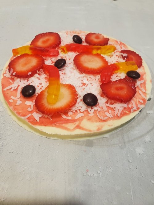 A colorful dessert pizza topped with strawberries, gummy worms, chocolate candies, and shredded coconut on a crust.