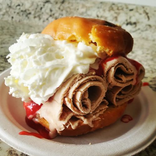 A donut topped with whipped cream and rolled chocolate ice cream, drizzled with strawberry sauce on a paper plate.