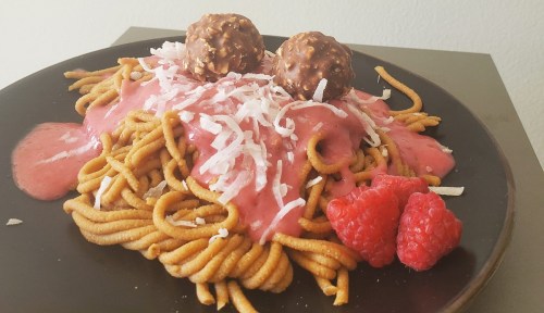A plate of spaghetti topped with pink sauce, shredded coconut, chocolate balls, and fresh raspberries.