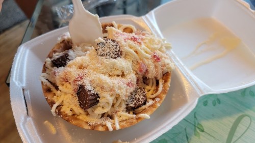 A dessert bowl filled with noodles, chocolate pieces, and shredded coconut, served in a takeout container.