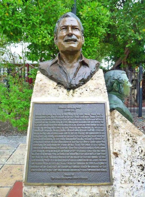 Bronze bust of Ernest Hemingway on a stone pedestal with an engraved plaque, surrounded by greenery.