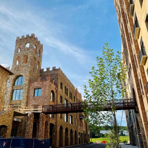 A brick building with a tower and a wooden bridge, surrounded by greenery and a clear blue sky.
