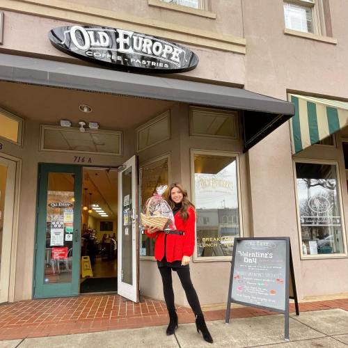 A woman in a red coat holds a gift basket outside Old Europe Coffee & Pastries, with a sign for Valentine's Day nearby.