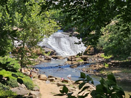 A serene waterfall cascades into a rocky stream, surrounded by lush greenery and people enjoying the natural scenery.