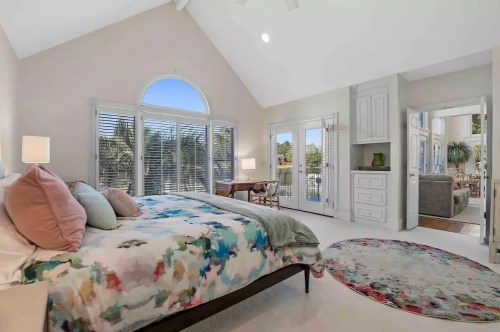 Bright bedroom with a colorful bedspread, large windows, and a cozy seating area visible through an open door.