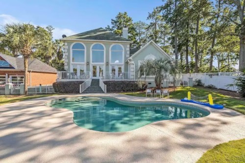 A spacious house with large windows overlooks a pool surrounded by palm trees and a well-maintained lawn.