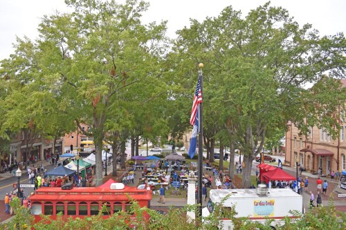 A bustling outdoor market scene with tents, people, and trees, featuring a red trolley and an American flag.