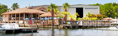 A marina with a restaurant, palm trees, and boats docked by a wooden pier, set against a clear blue sky.