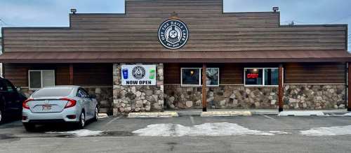 A rustic building with a stone facade and a sign reading "NOW OPEN," featuring an open sign and parked cars outside.