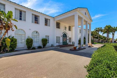 A white building with columns and palm trees, featuring a clear blue sky and a landscaped entrance.
