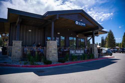 A rustic restaurant exterior with large windows, stone pillars, and outdoor seating, set against a blue sky.