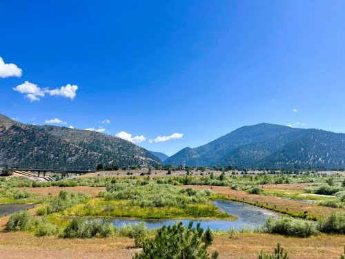 A scenic landscape featuring a river, lush greenery, and mountains under a clear blue sky.