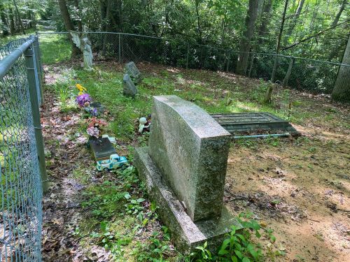 A fenced cemetery with a gravestone, flowers, and small decorations among trees and greenery.