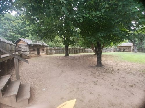 A grassy area surrounded by trees and wooden cabins, with a wooden fence in the background.