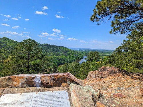 Scenic view of a river winding through green hills under a blue sky, with rocky foreground and trees.