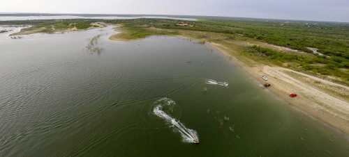 Aerial view of a calm lake with a boat creating ripples, surrounded by green land and a sandy shore.