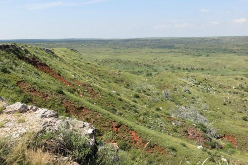 A panoramic view of rolling green hills with patches of red soil under a clear blue sky.