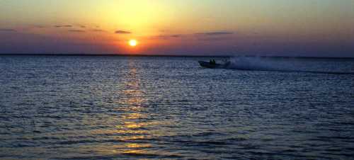 A boat speeds across a calm lake at sunset, with vibrant orange and purple hues in the sky.