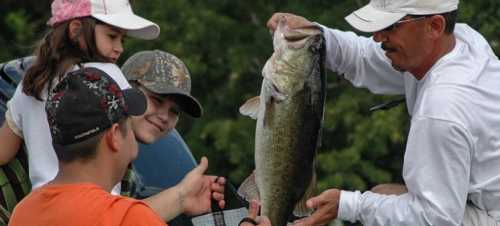 A man shows a large fish to two children while a girl watches, all enjoying a day outdoors.