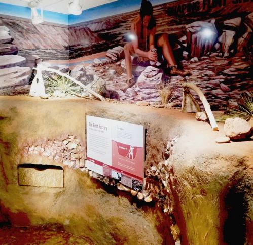 A museum exhibit featuring a rocky landscape, a mural of a person crafting tools, and an informational display.