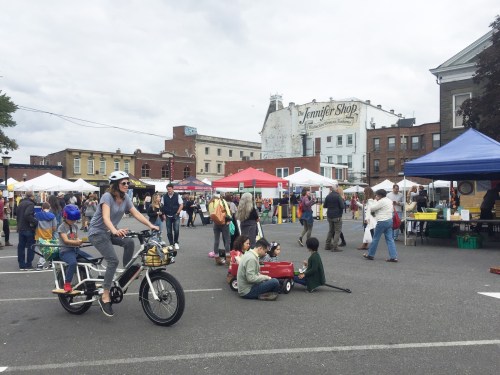 A bustling outdoor market scene with people shopping, tents set up, and a cyclist passing by with children.