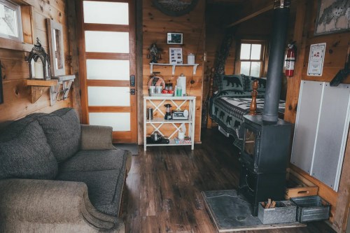 Cozy wooden interior of a cabin featuring a sofa, wood stove, and shelves with decor and essentials.