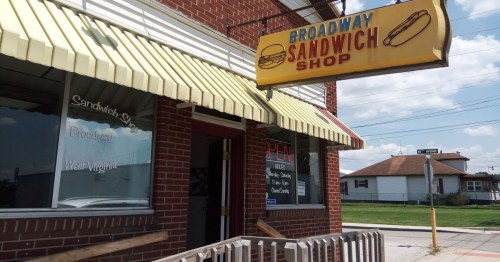 Exterior of Broadway Sandwich Shop with a yellow awning and a sign featuring a hot dog, under a blue sky.