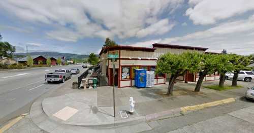 Street view of a corner building with trees, parked cars, and a blue vending machine on a sunny day.