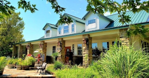 A charming house with a stone facade, green roof, and a welcoming porch surrounded by lush greenery and plants.