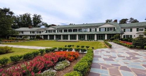 A large, elegant house surrounded by colorful flower beds and a well-maintained lawn under a cloudy sky.