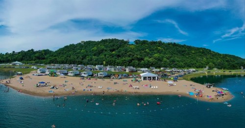 Aerial view of a beach with people swimming, surrounded by green hills and camper trailers under a blue sky.