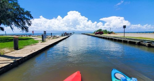 A serene waterway leads to a beach, with kayaks in the foreground and fluffy clouds in a blue sky above.