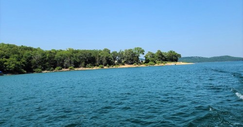 A serene view of a calm blue lake with a sandy shore and lush green trees under a clear blue sky.