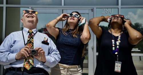 Three people wearing eclipse glasses look up, smiling, in front of a building on a sunny day.