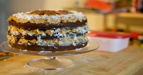 A three-layer chocolate cake topped with coconut and pecans, displayed on a glass cake stand.