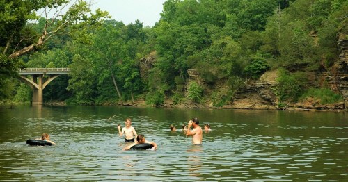 A group of children playing in a river, surrounded by lush greenery and a bridge in the background.