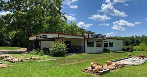 A mining shop with a metal roof, surrounded by grass and trees, under a blue sky with fluffy clouds.