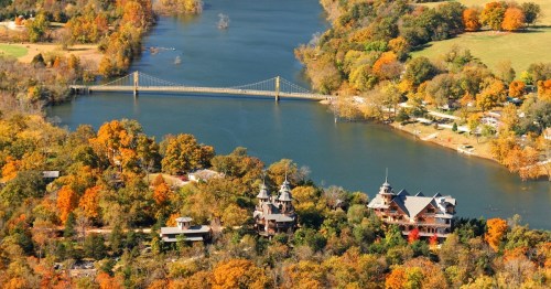 Aerial view of a river with a bridge, surrounded by colorful autumn trees and a charming house on the shore.