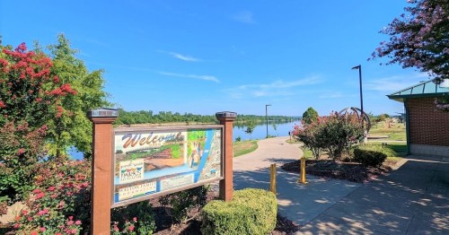 A scenic park entrance sign by a calm lake, surrounded by greenery and colorful flowers under a clear blue sky.