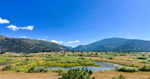 A scenic landscape featuring a river, lush greenery, and mountains under a clear blue sky.