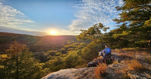 A person sits on a rocky ledge, overlooking a lush valley at sunset, surrounded by trees and vibrant sky colors.