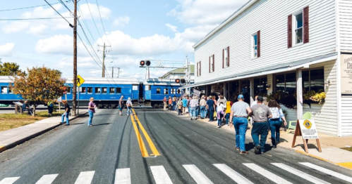 A crowd walks along a street as a blue train passes by a white building with windows and a sign.