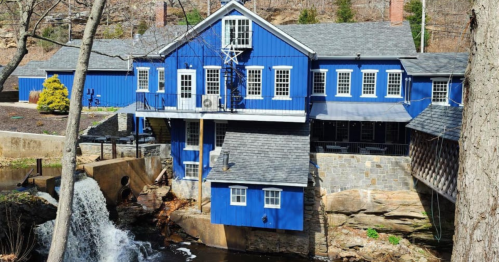 A blue building by a stream, featuring large windows and a waterfall in the foreground, surrounded by trees.