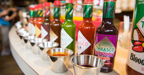 A row of various Tabasco sauce bottles with small metal cups in front, showcasing different flavors and colors.