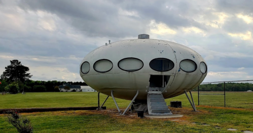 A futuristic, oval-shaped building on stilts, set in a grassy field under a cloudy sky.