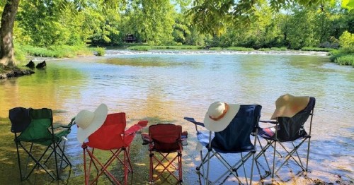 Five camping chairs in a row by a calm river, surrounded by lush greenery and sunlight filtering through the trees.