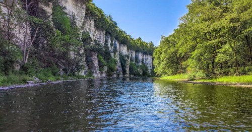 A serene river flows between lush green cliffs under a clear blue sky, surrounded by vibrant trees.