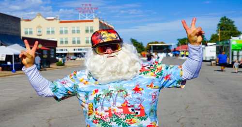 A person dressed as Santa in a colorful Hawaiian shirt and sunglasses, posing with peace signs in a festive street setting.