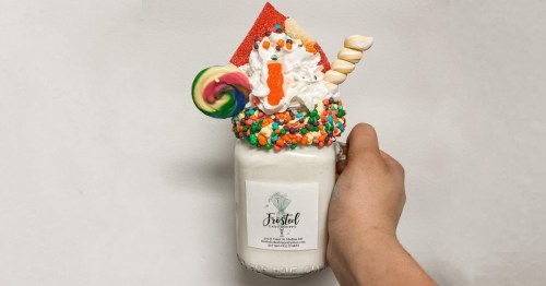 A hand holding a decorated milkshake in a jar, topped with whipped cream, candy, and colorful sprinkles.