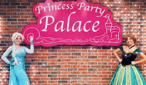 Two women in princess costumes stand beside a pink sign that reads "Princess Party Palace," against a brick wall.